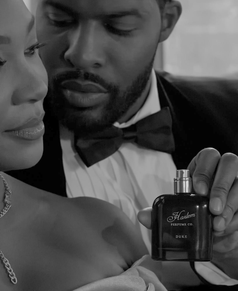 A black perfume bottle labeled 'Harlem Perfumes Co. DUKE' is held by a man in a tuxedo while a woman in a strapless gown gazes at him.