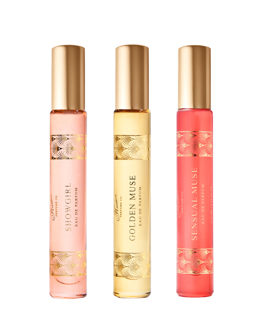 Three perfume travel spays with gold caps on a white background: a pale pink bottle labeled SHOWGIRL, a pale yellow bottle labeled GOLDEN MUSE, and a coral pink bottle labeled SENSUAL MUSE.