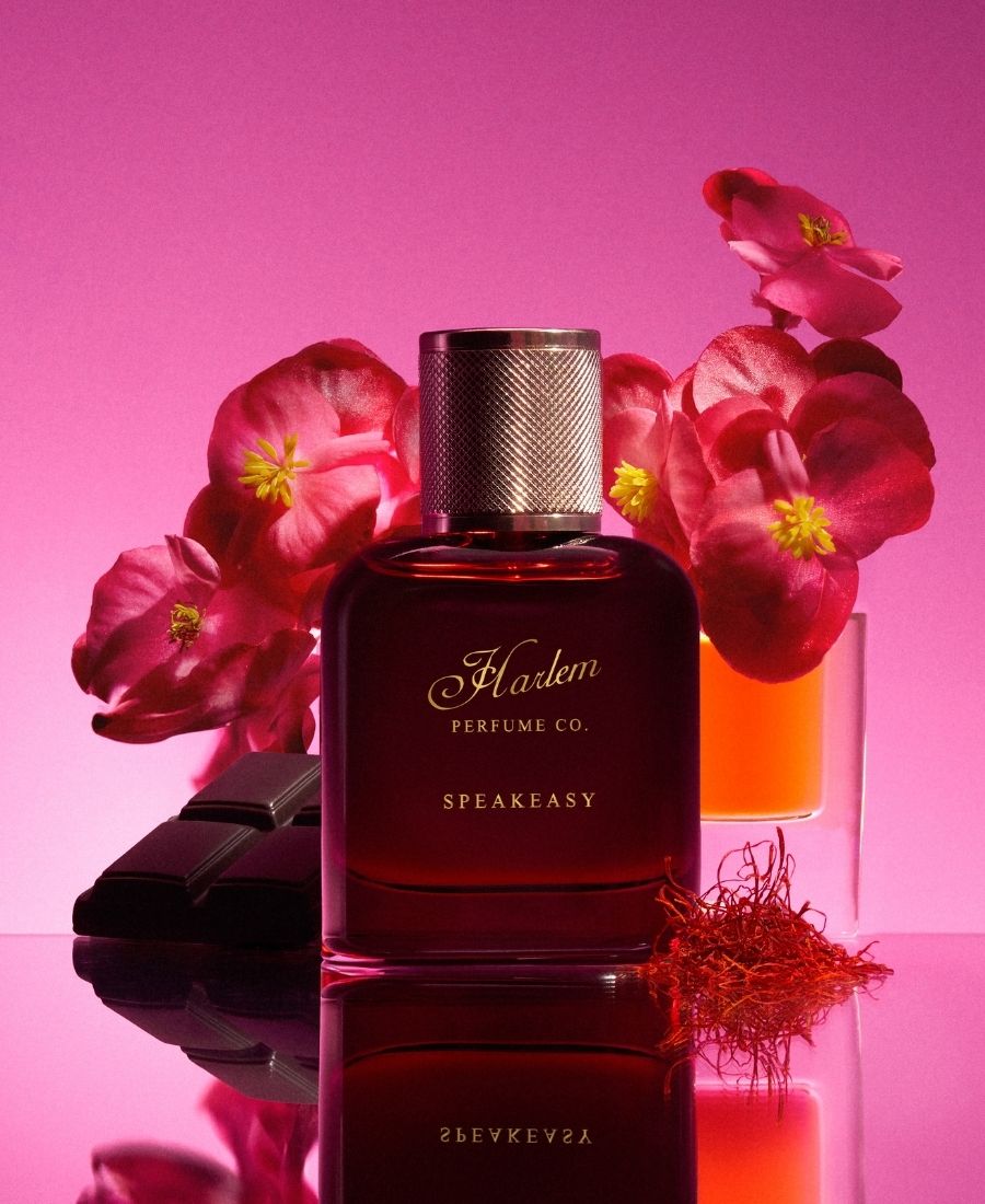 This is an image of the Speakeasy 50ml Eau De Parfum bottle surrounded by white and magenta flowers against a pnk background.