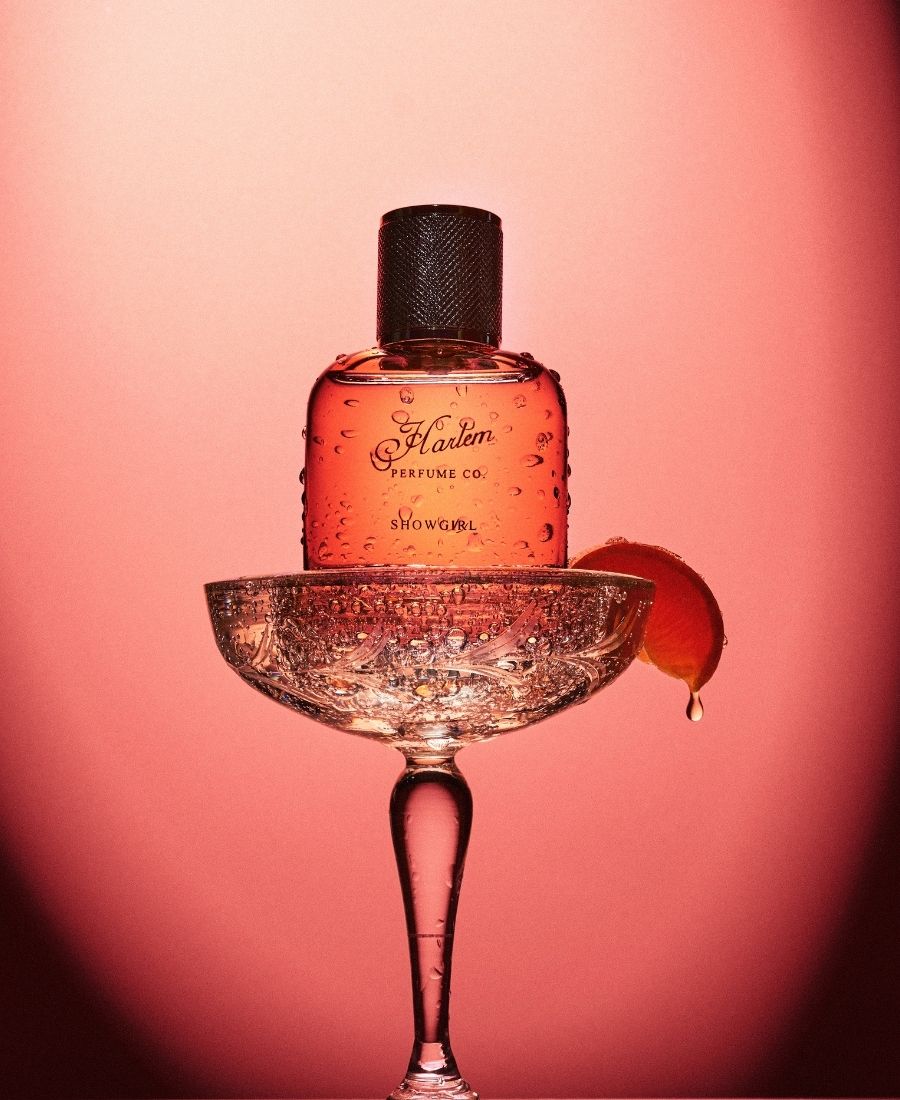 This is a lifestyle image of the Showgirl in a vintage champagne glass with a mandarin slice hanging off the side. The peachy pink bottle has water droplets and the background image is pink.