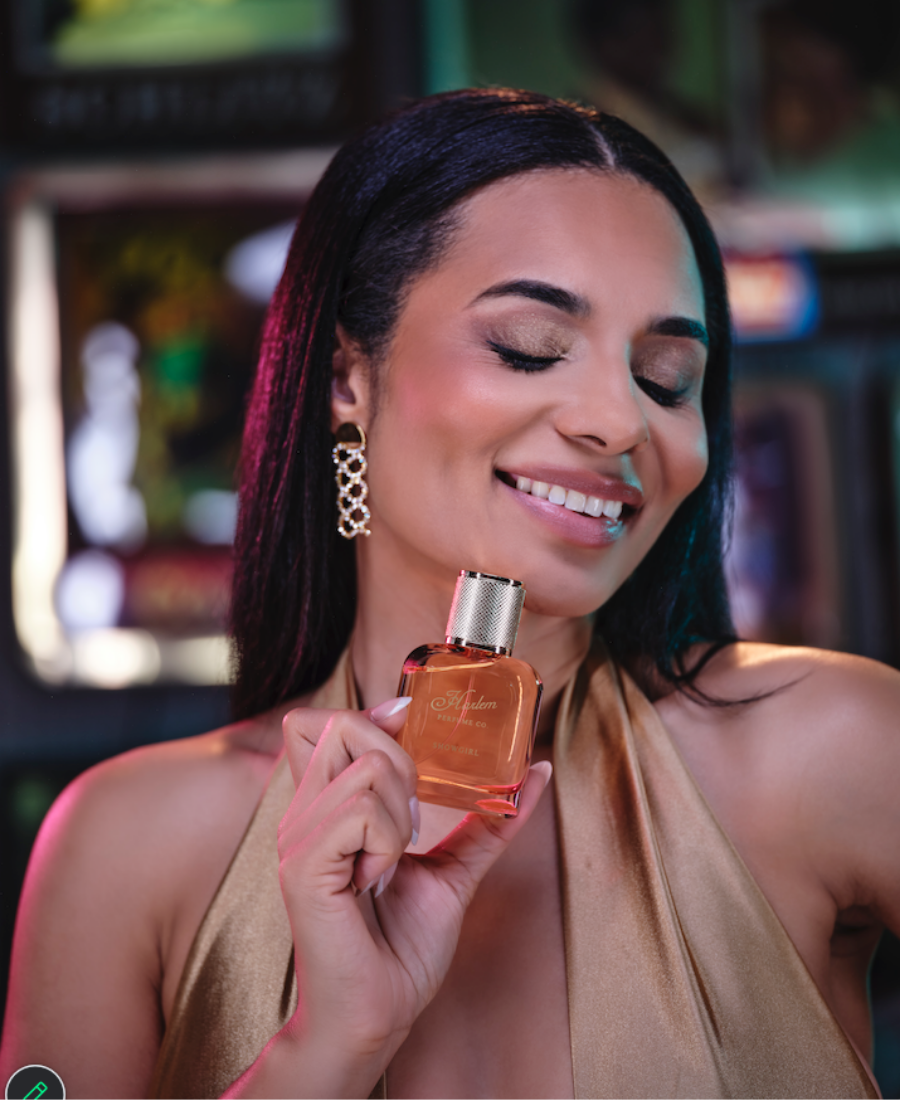 A woman with dark hair holds a bottle of orange-amber fragrance near her face. She is wearing a champagne-colored halter top and gold statement earrings, with a softly blurred warm-toned indoor background.