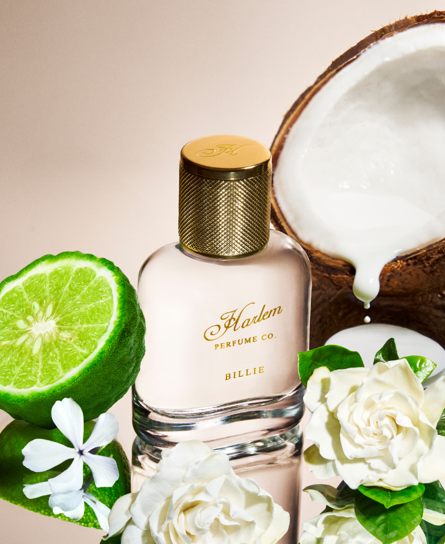 This is an image of our Billie eau de Parfum bottle in a lifestyle setting surrounded by bergamot,  The bottle is clear with gold letters that say Harlem Perfume Co and the word Billie as the name of the fragrance, coconut milk and gardenia.