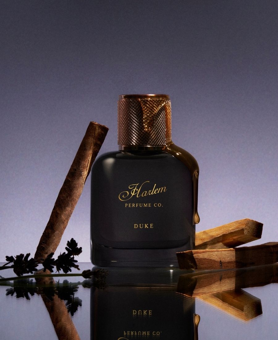 This is an image of our Duke black perfume bottle with  the 'Harlem Perfume Co.' logo positioned on a reflective surface with palo santo, lavender, and honey dripping and a cigar with tobacco leaning next to it.