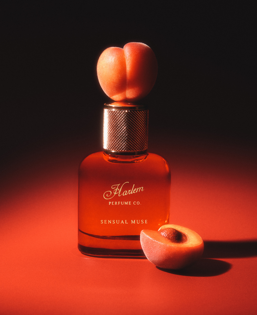 A perfume bottle labeled 'Harlem Perfume Co. Sensual Muse' with an apricot shown on a dark red and black background with a sliced peach in front.