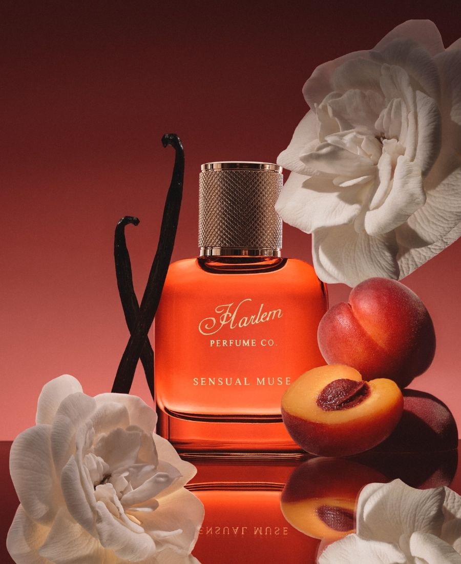 Orange perfume bottle labeled 'Harlem Perfume Co Sensual Muse' with vanilla bean, apricot, and white flowers on a reflective surface.